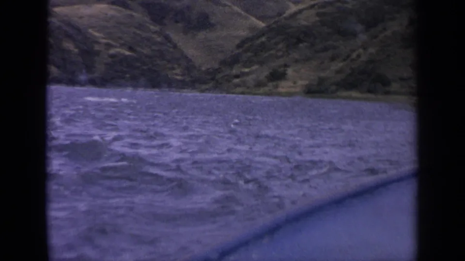 Boat-level 1963 Idaho lake view with wind-driven waves, a visible rail or hull edge, and rugged hills in the background.