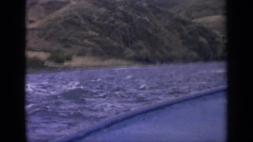1963 Idaho boat-window view of rough blue river water, whitecaps, and a distant green-brown hillside shoreline.