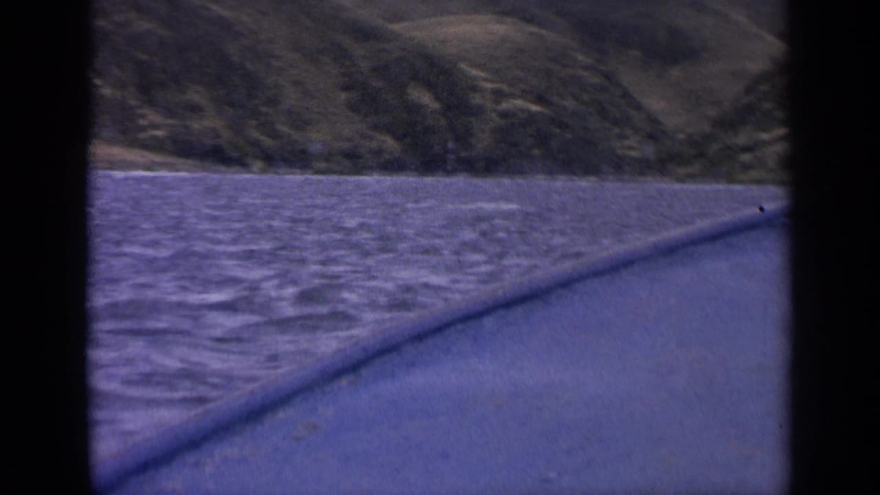 1963 Idaho river scene framed by a boat window, with rippled water, a low wooded shoreline, and hills beyond.