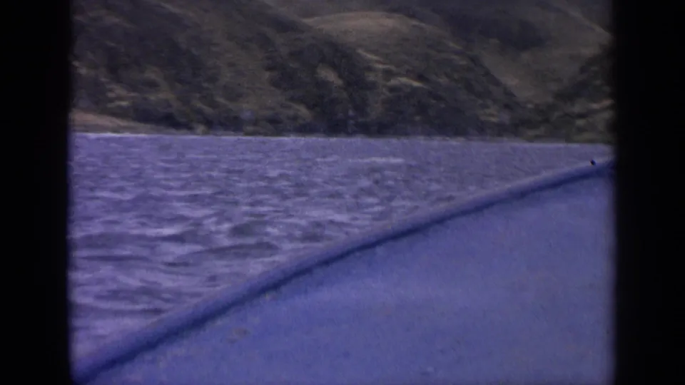 1963 Idaho river scene framed by a boat window, with rippled water, a low wooded shoreline, and hills beyond.