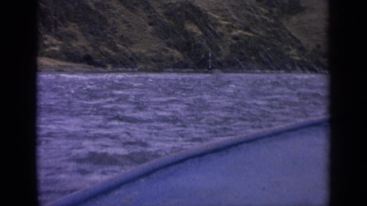 1963 Idaho boat POV over choppy river water toward green-brown hills, with window framing visible in the foreground.