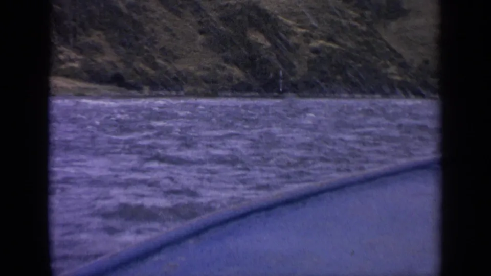 1963 Idaho boat POV over choppy river water toward green-brown hills, with window framing visible in the foreground.