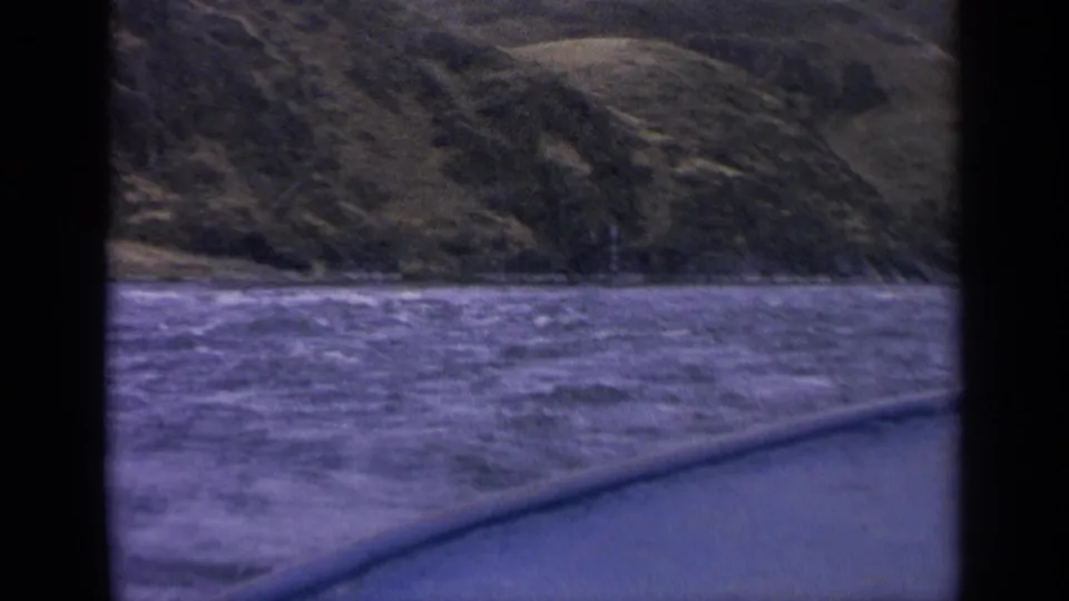 1963 Idaho 8mm boat view across choppy lake water toward a rugged hillside shoreline, framed by dark film or window edges.