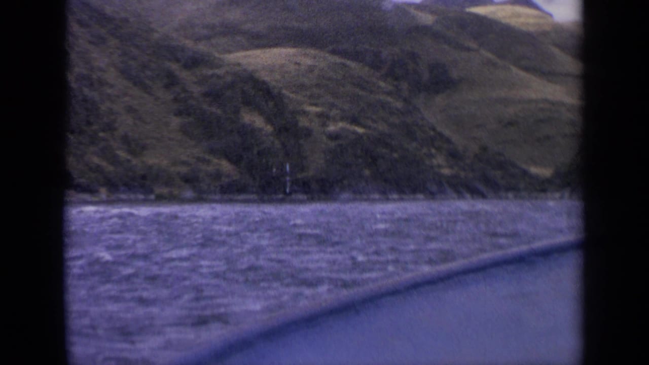 1963 Idaho home-movie frame with rough lake water in front of a sloping shoreline, seen from a moving boat with dark edge borders.