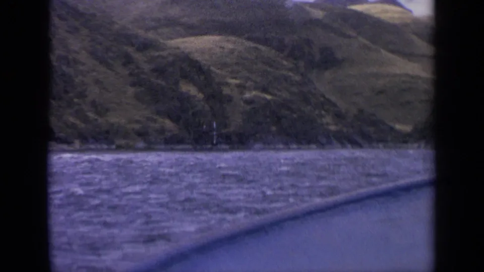 1963 Idaho home-movie frame with rough lake water in front of a sloping shoreline, seen from a moving boat with dark edge borders.