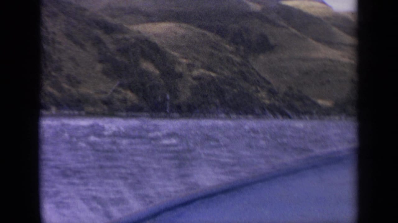 1963 Idaho 8mm view across purple-tinted lake water toward rolling hills, with dark frame edges and a boat edge in the foreground.