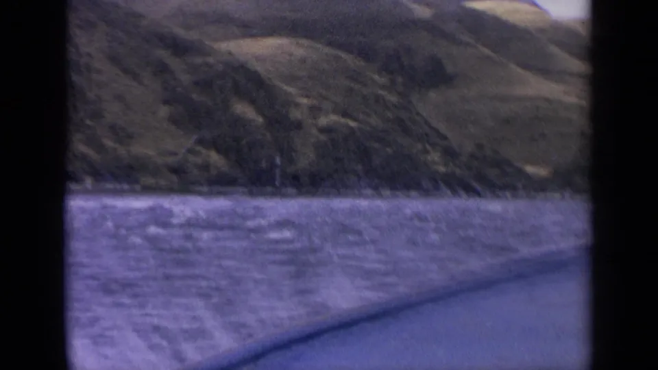 1963 Idaho 8mm view across purple-tinted lake water toward rolling hills, with dark frame edges and a boat edge in the foreground.