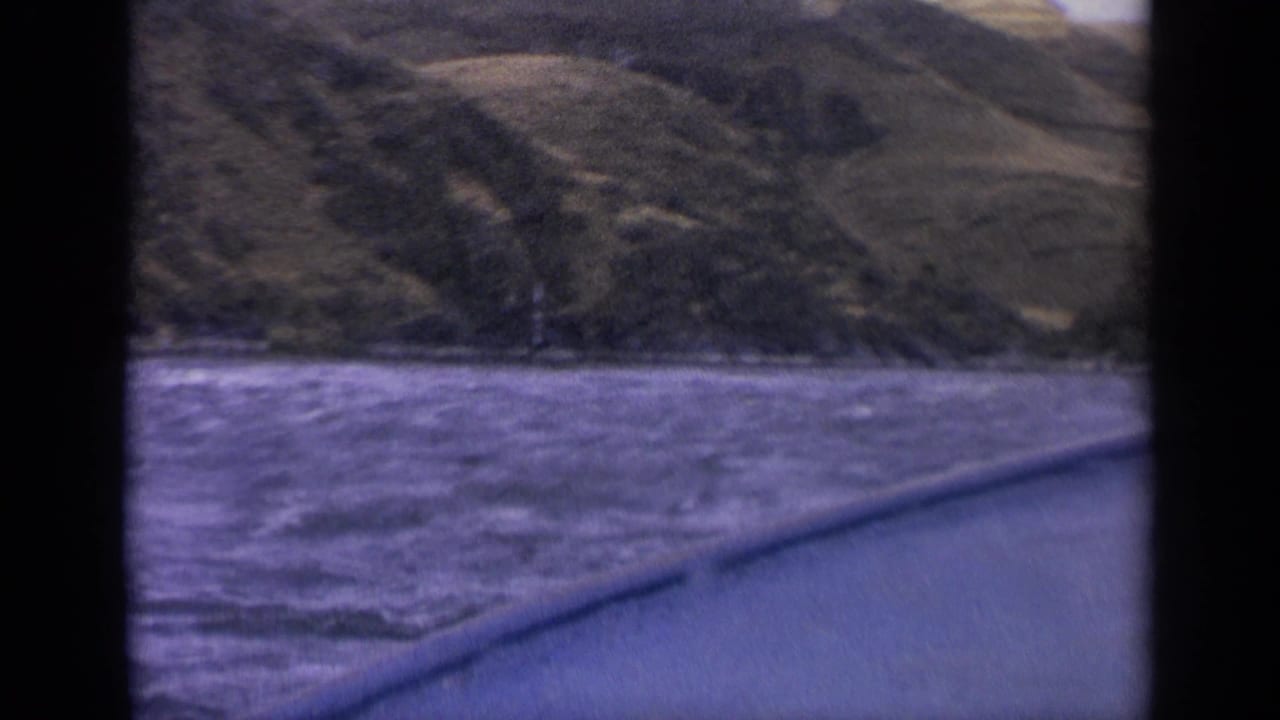 1963 Idaho 8mm frame from a boat, showing rough river water, a dark gunwale, and green-brown mountain slopes beyond the shoreline.