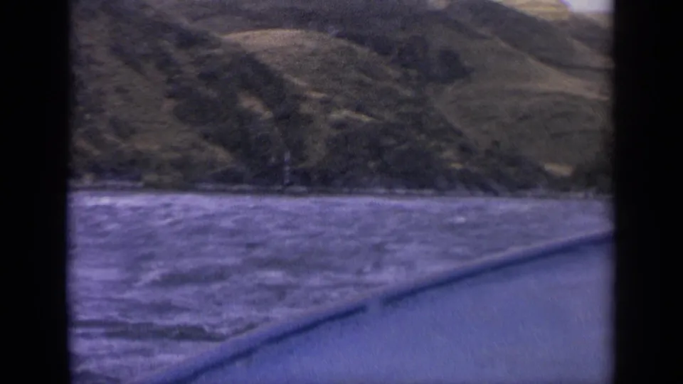 1963 Idaho 8mm frame from a boat, showing rough river water, a dark gunwale, and green-brown mountain slopes beyond the shoreline.