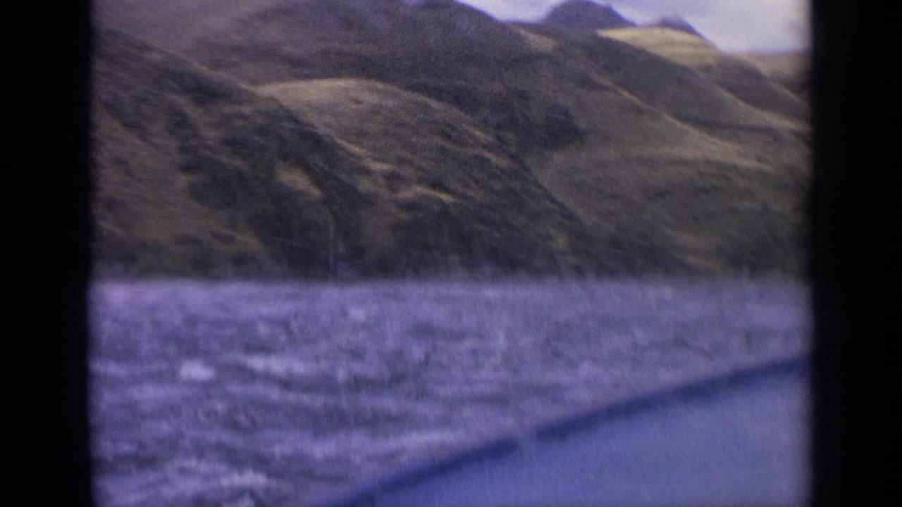 1963 Idaho home-movie frame with purple-tinged river water, a slanted boat edge, and steep green hills rising from the far bank.