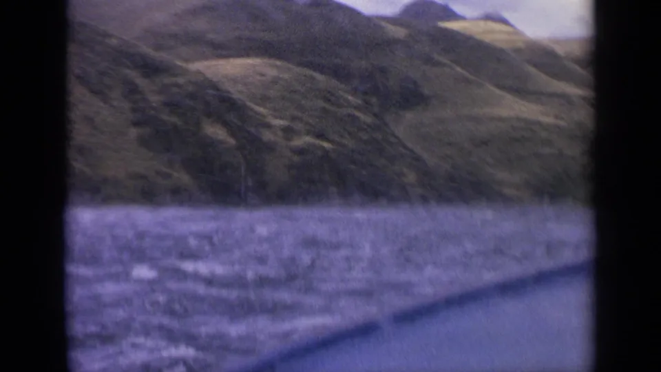 1963 Idaho home-movie frame with purple-tinged river water, a slanted boat edge, and steep green hills rising from the far bank.