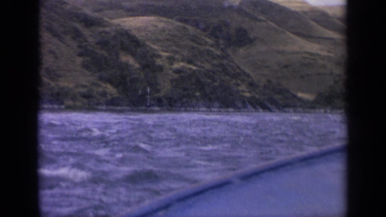 1963 Idaho 8mm still looking from a boat across rippling river water toward a steep, grassy bank and mountain slope.