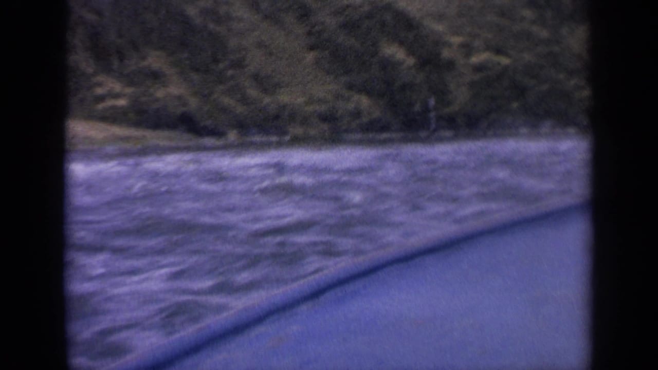 Final 1963 Idaho frame with rough river water beside the boat and a dark, steep shoreline filling the background.