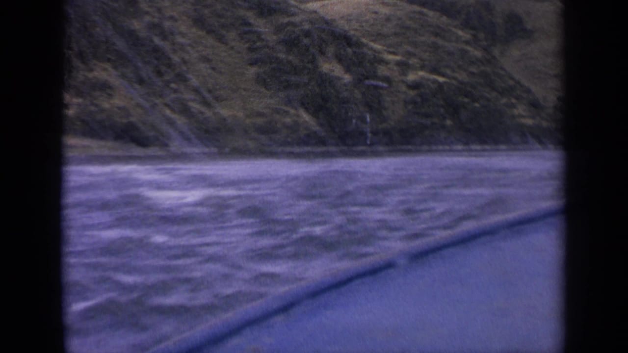 1963 Idaho 8mm frame showing turbulent river water from a moving boat, with a sloped grassy hillside beyond.