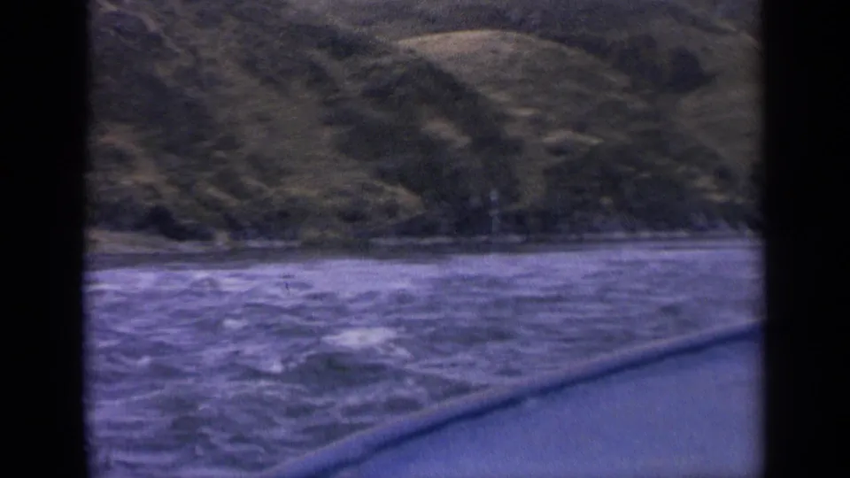 1963 Idaho home-movie frame showing textured river water, a low shoreline, and a grassy hillside from a moving boat.