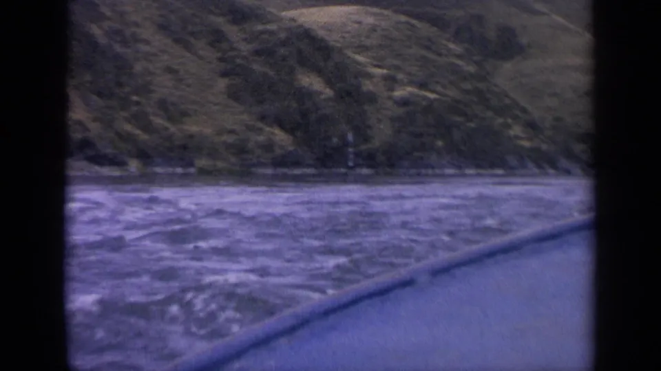 Final 8mm frame of a moving boat view across foamy Idaho river water toward a grassy hillside shoreline.