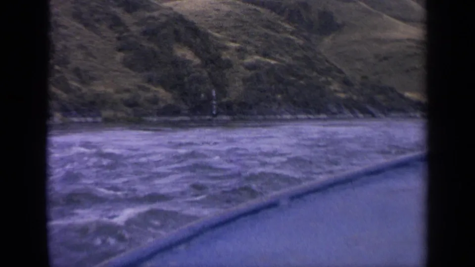 1963 Idaho 8mm view from a boat, showing churning water, a blue foreground edge, and a grassy hillside shoreline.