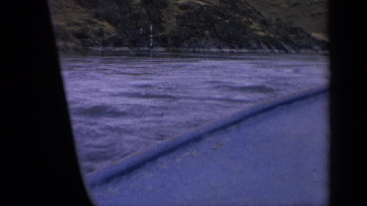Boat passenger view in 1963 Idaho, with rough blue-purple water, shoreline brush, and dark vertical framing from the filming position.