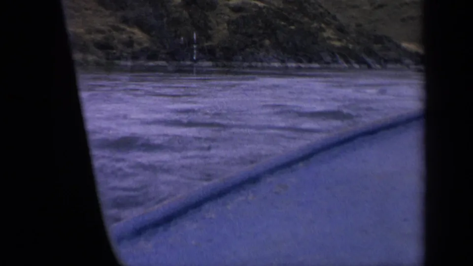 Boat passenger view in 1963 Idaho, with rough blue-purple water, shoreline brush, and dark vertical framing from the filming position.