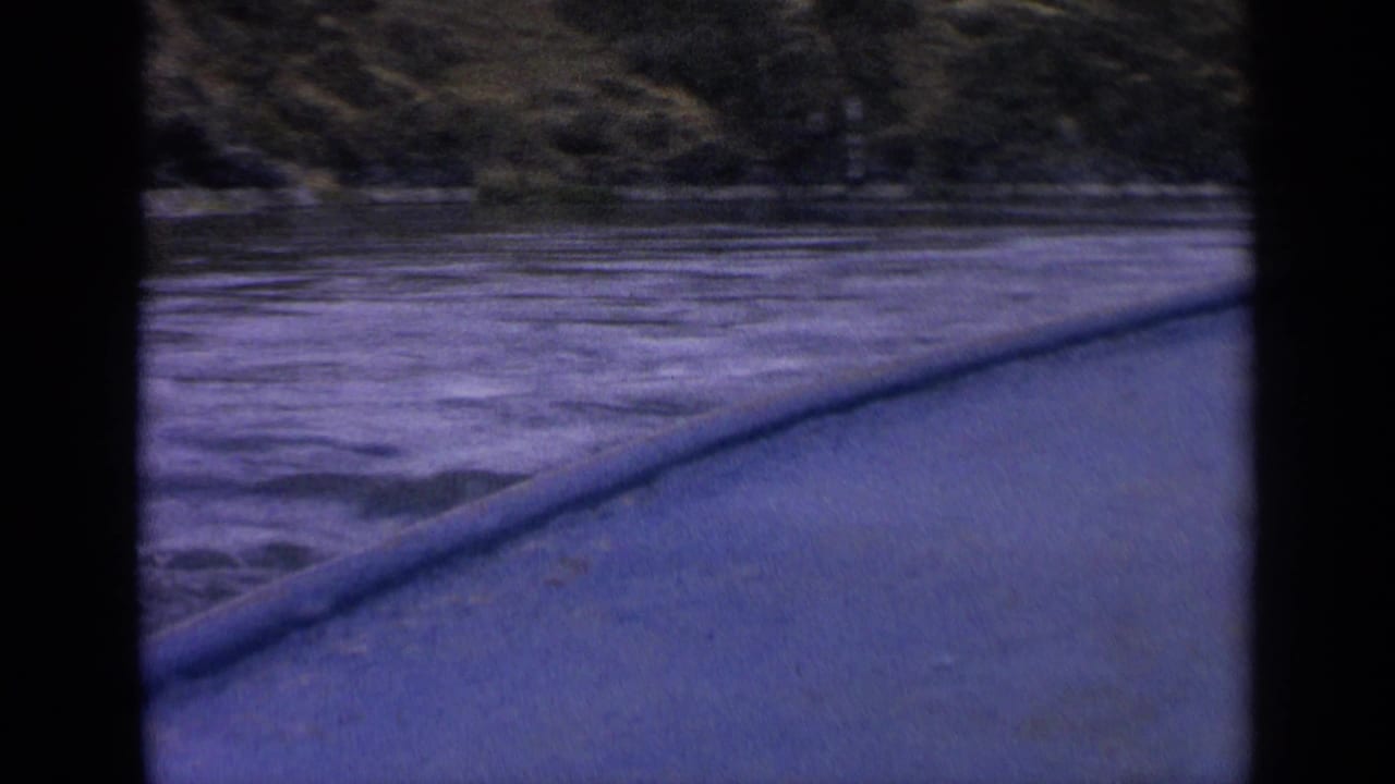 1963 Idaho home-movie frame showing the side of a boat moving past rippled water and a shadowed grassy shore.