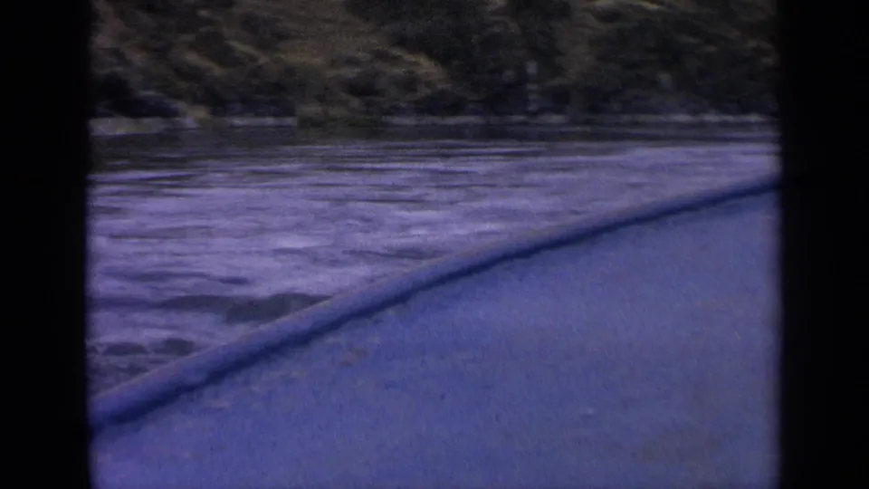 1963 Idaho home-movie frame showing the side of a boat moving past rippled water and a shadowed grassy shore.