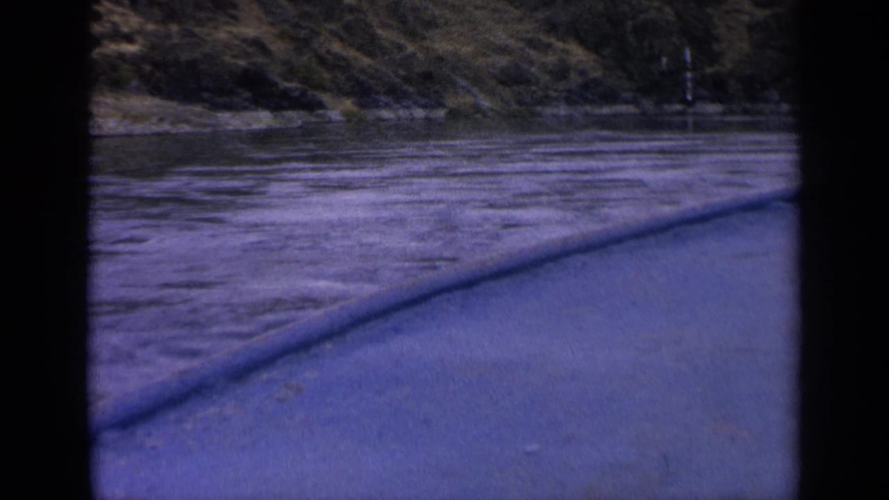 End frame from a 1963 Idaho 8mm boat trip, with choppy water, blue foreground boat edge, and rugged grassy shore.