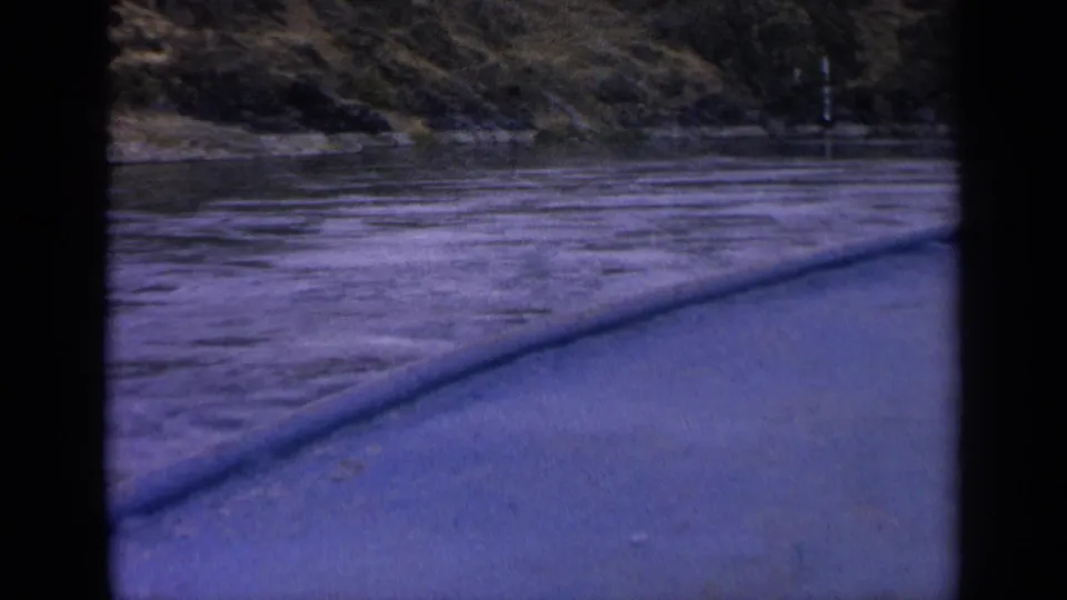 End frame from a 1963 Idaho 8mm boat trip, with choppy water, blue foreground boat edge, and rugged grassy shore.