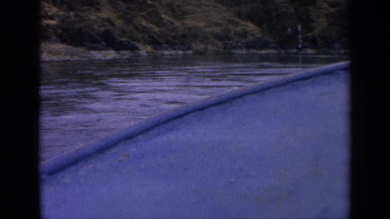 1963 Idaho 8mm boat-view frame showing rippled river water, a low bow or rail, and a wooded hillside beyond the shore.