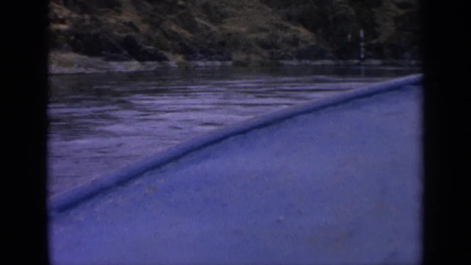 1963 Idaho 8mm boat-view frame showing rippled river water, a low bow or rail, and a wooded hillside beyond the shore.