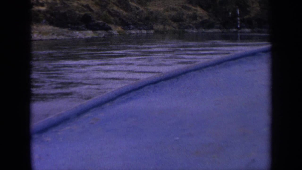 Boat perspective on an Idaho river in 1963, with the blue-toned bow or rail leading toward water, shoreline, and dark hillside trees.