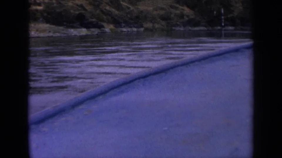 Boat perspective on an Idaho river in 1963, with the blue-toned bow or rail leading toward water, shoreline, and dark hillside trees.