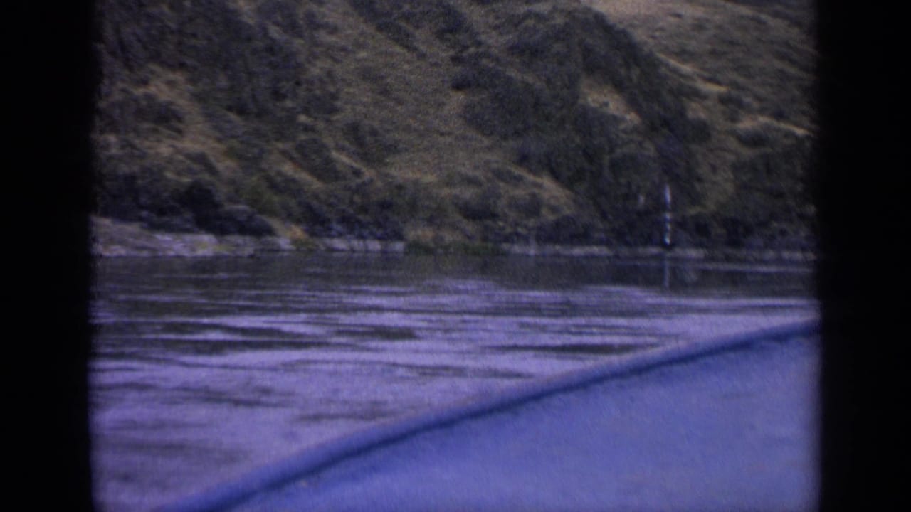 1963 Idaho home-movie frame from a boat, showing rippled river water, wooded banks, and a hillside under soft, aged 8mm color.