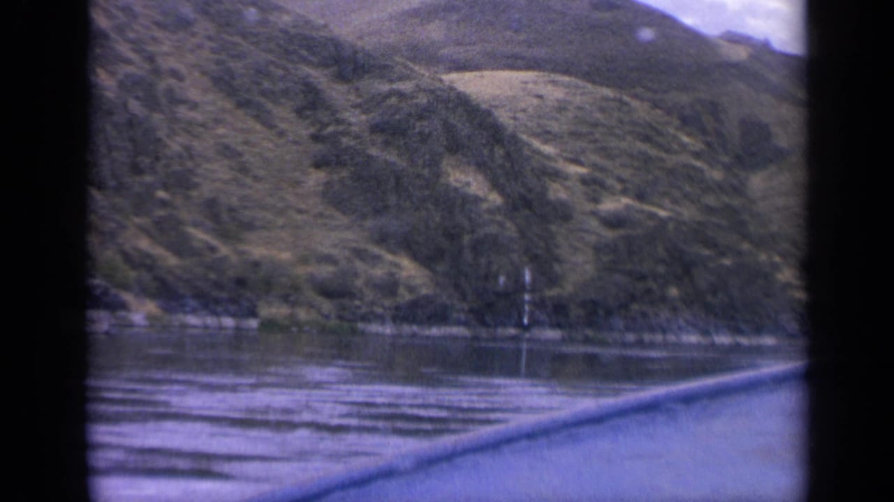 Final 1963 Idaho boat-view frame looking over calm water to open grassy hills, scattered trees, and distant slopes.