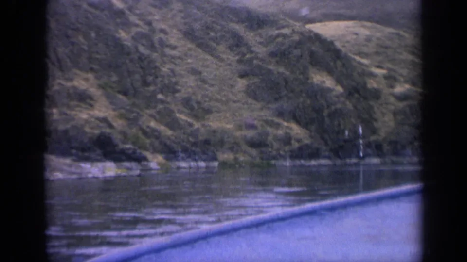 1963 Idaho home-movie view of lake water, steep tree-dotted hills, rocky shore, and a blue boat or window edge in front.
