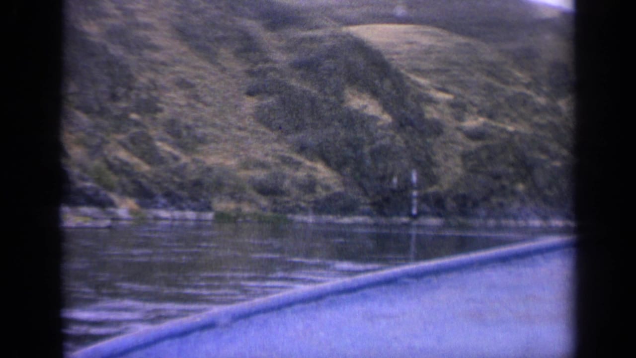 1963 Idaho 8mm still with lake water, rocky shoreline, sloping mountains, scattered trees, and heavy dark frame borders.