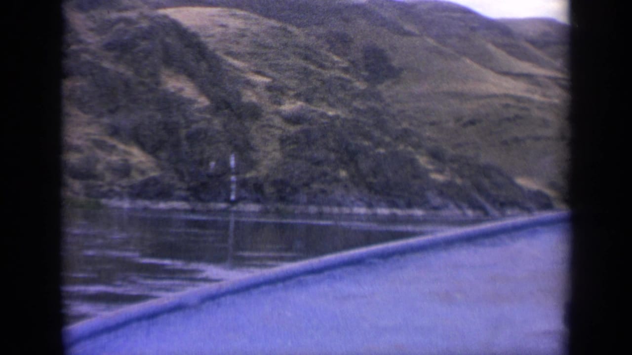 Final 1963 Idaho frame showing a calm lake, steep mountain terrain, sparse trees, and the boat or window edge at lower right.