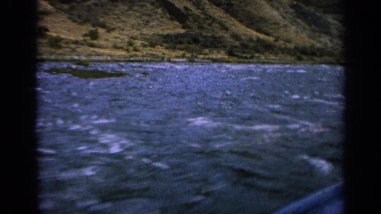 1963 Idaho 8mm frame showing whitecapped river water, a rocky bank, and a grassy hillside beyond.