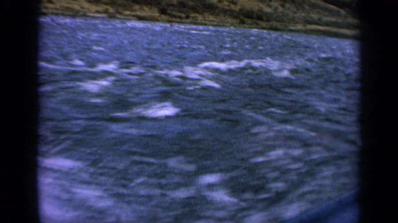 Boat-level 8mm view of rough Idaho river water with a grassy mountain slope beyond.