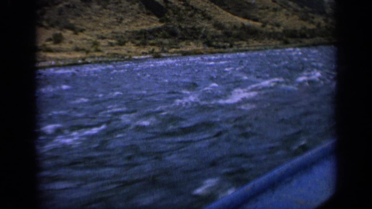 1963 Idaho 8mm frame showing a first-person view over choppy river water from a moving boat, with a grassy bank in the distance.