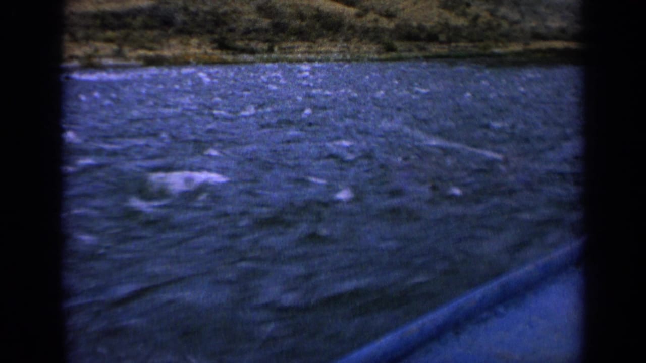 1963 home-movie frame of blue river water, scattered white wake, and a blurred earth-toned bank in Idaho.