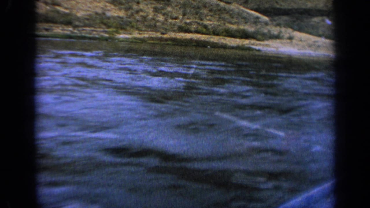 1963 Idaho home-movie frame showing rippled river water, a low bank, and hillside from a moving boat perspective.