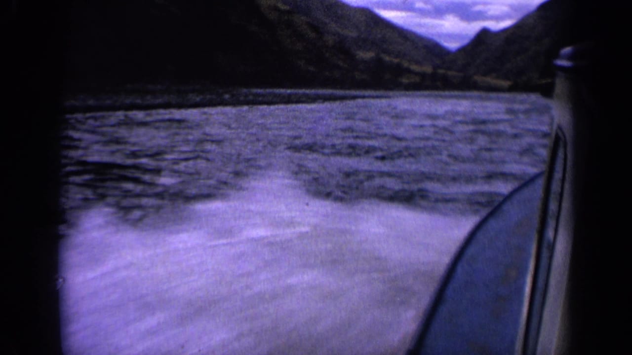 1963 Idaho 8mm frame from a moving boat, showing a foamy wake, blue water, mountain shoreline, and cloudy sky.
