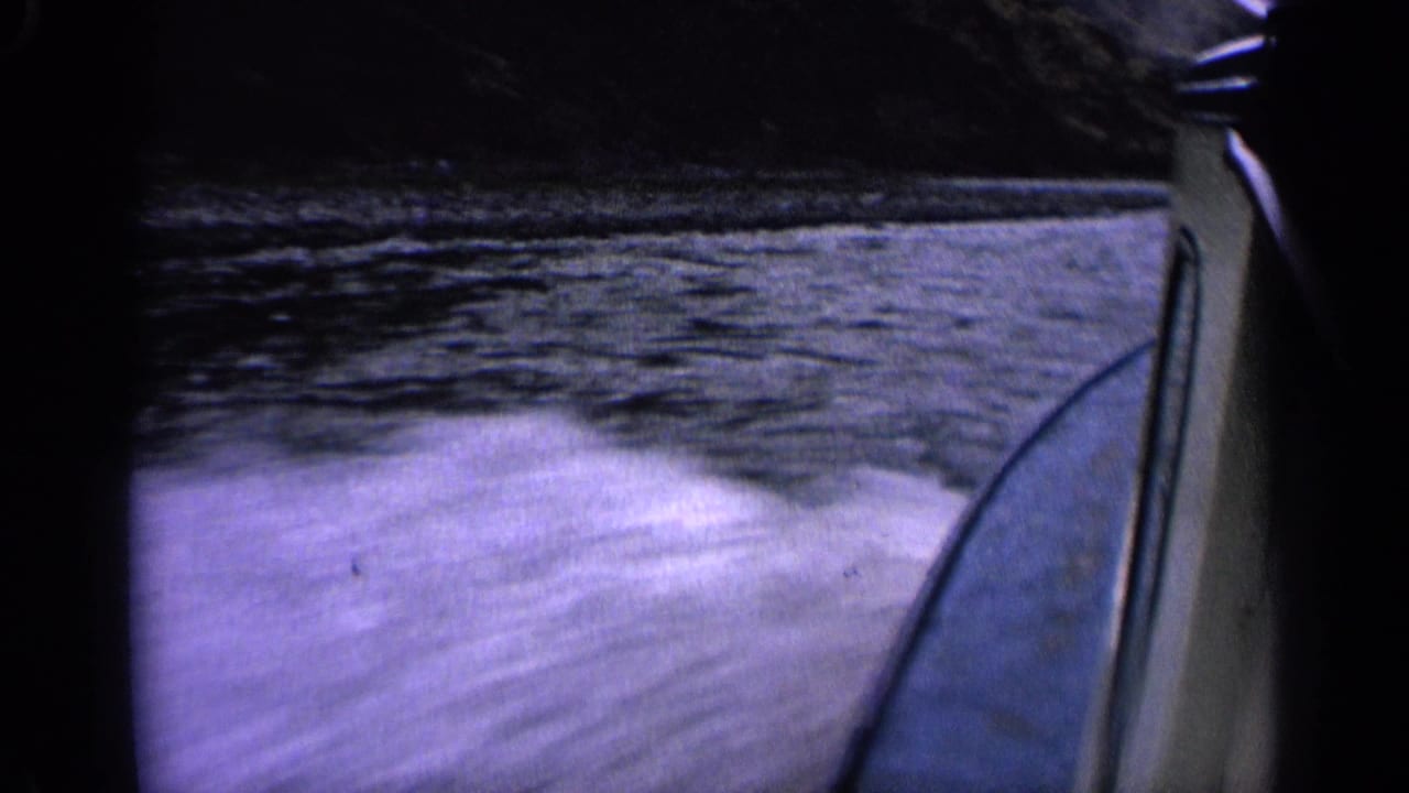 1963 Idaho home-movie frame of a boat wake rushing past, with blue water, mountain shoreline, and the boat edge visible.
