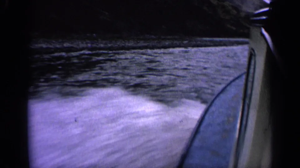 1963 Idaho home-movie frame of a boat wake rushing past, with blue water, mountain shoreline, and the boat edge visible.
