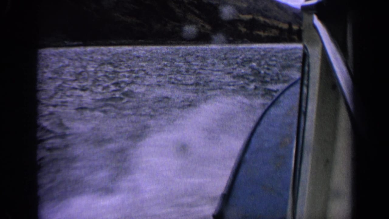1963 Idaho home-movie frame with a boat edge cutting past churning wake on a lake near a wooded shoreline.