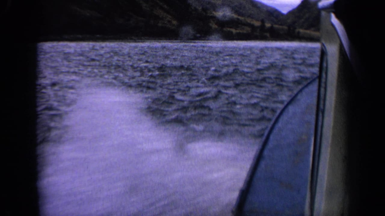 1963 Idaho 8mm frame from a moving boat, showing churned wake, rippled water, the hull edge, and dark wooded hills in the distance.
