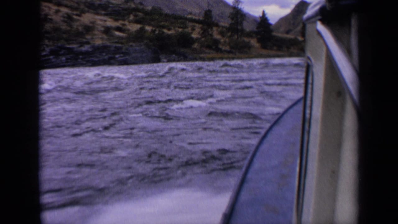 8mm passenger-side boating frame from 1963 Idaho, showing foamy wake, rough water, the boat edge, trees, and hills along the shore.