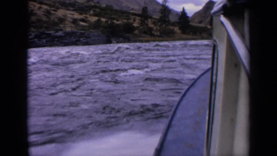 8mm passenger-side boating frame from 1963 Idaho, showing foamy wake, rough water, the boat edge, trees, and hills along the shore.