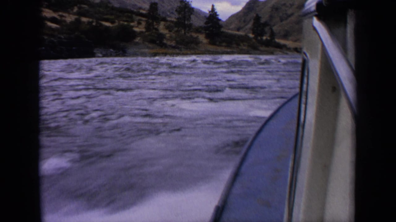 1963 Idaho home-movie frame from a speedboat, with blurred wake in front, the hull edge at right, and wooded hills across the water.