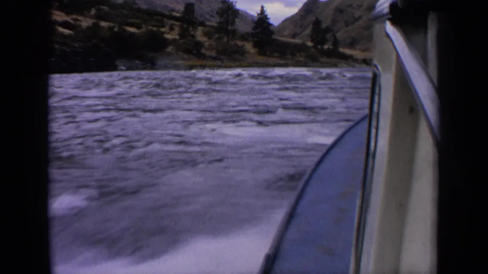 1963 Idaho home-movie frame from a speedboat, with blurred wake in front, the hull edge at right, and wooded hills across the water.